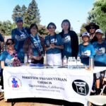 A group of people, adults and children, pose outdoors behind a table with a Parkview Presbyterian Church banner. Most wear blue Ocean Walk shirts and leis. Water bottles and pamphlets are on the table. Trees and a fence are in the background.