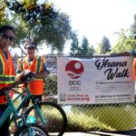Three people in safety vests stand by a bike, smiling, next to a fence with a banner that reads Ohana Walk and gives event details for ACC Senior Services in Sacramento, California. Trees and sunlight are in the background.