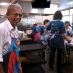 A smiling man wearing a white shirt and a Superman apron stands in the foreground of a busy kitchen, while several people in blue shirts prepare food at tables behind him.