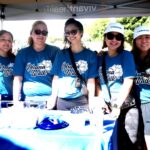 Five women wearing matching blue Ohana Walk t-shirts smile at a table under a tent at an outdoor event. Some wear sunglasses and hats, and there are various items on the table in front of them.