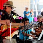 A group of older adults play guitars, keyboard, and sing on stage at an outdoor event. Some audience members watch and take photos. The setting is festive, with red curtains and bright lighting.