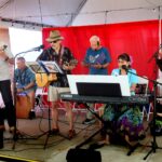 A band of six older adults performing on a small stage under a canopy. Two play ukuleles, one plays guitar, one plays keyboard, one plays a cajón drum, and one reads from a folder. They are dressed in colorful, casual clothing.