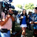 A cameraman films a smiling woman in a blue shirt and white skirt as she talks and laughs with a man holding a bowl at an outdoor event, surrounded by people and tents on a sunny day.