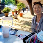 A woman with a painted face smiles at an outdoor table covered with art supplies, drinks, and snacks. Potted plants and other people are visible in the background on a sunny day.