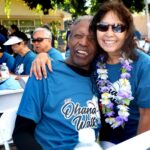Two people smiling and posing together at an outdoor event, both wearing blue Ohana Walk T-shirts. The woman wears sunglasses and a flower lei. Several people are seated at tables in the background.