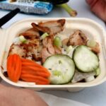 A takeout container with grilled chicken, sliced carrots, cucumber, chopped green onions, and rice. A persons hand is holding the container, and a wrapped granola bar is in the background.