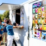 Four people stand in line at a white food truck, ordering drinks. A colorful menu with various bubble tea options and images is displayed on the side of the truck. Trees and grass are visible in the background.