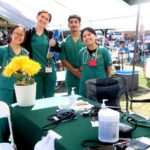 Four smiling healthcare workers in green scrubs stand outdoors by a table with medical supplies and potted yellow flowers. Behind them, many people sit under a large canopy at a community event.