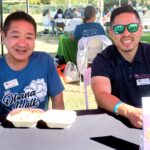 Two people sit at a table outdoors, smiling. One wears a blue “Ohana Walk” shirt, the other wears sunglasses and a dark polo. They have food containers and drinks in front of them. Other people and tables are visible in the background.