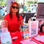 A woman with long red hair and sunglasses sits at an outdoor booth with AARP brochures, a raffle sign, and an emergency backpack prize displayed on the table. Other people and booths are visible in the background.