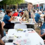 A Boy Scout in uniform carries a tray while standing by a table at a crowded outdoor event, where people are eating and socializing under tents.