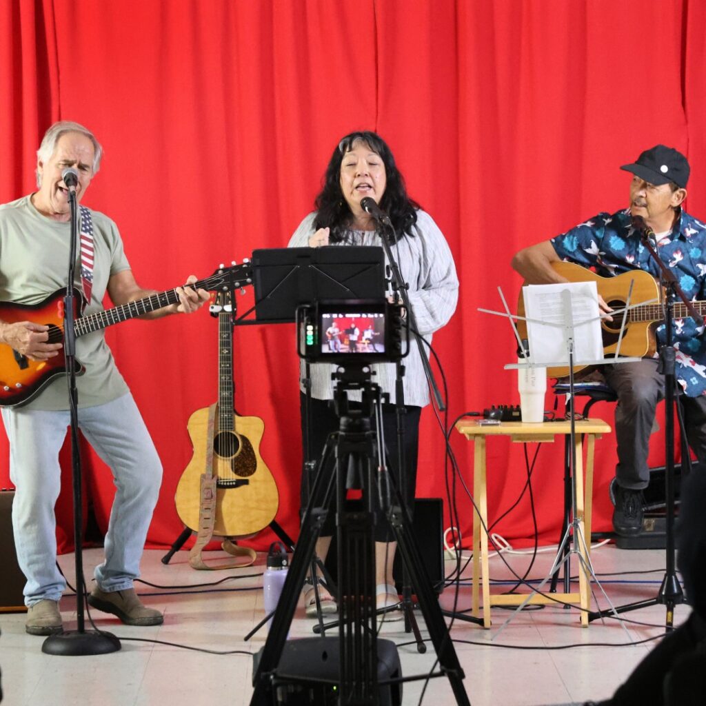 Three musicians perform on stage in front of a red curtain: a man playing electric guitar, a woman singing into a microphone, and another man playing acoustic guitar. A camera on a tripod records them.