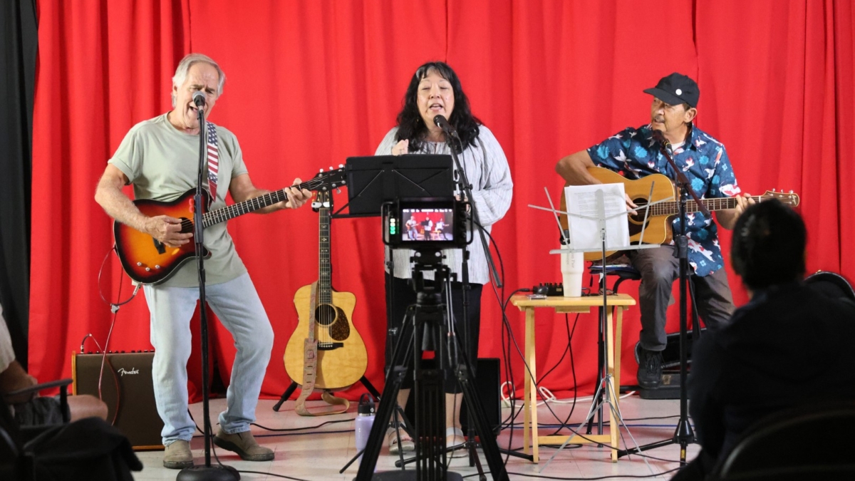Three musicians perform on stage in front of a red curtain: a man playing electric guitar, a woman singing into a microphone, and another man playing acoustic guitar. A camera on a tripod records them.