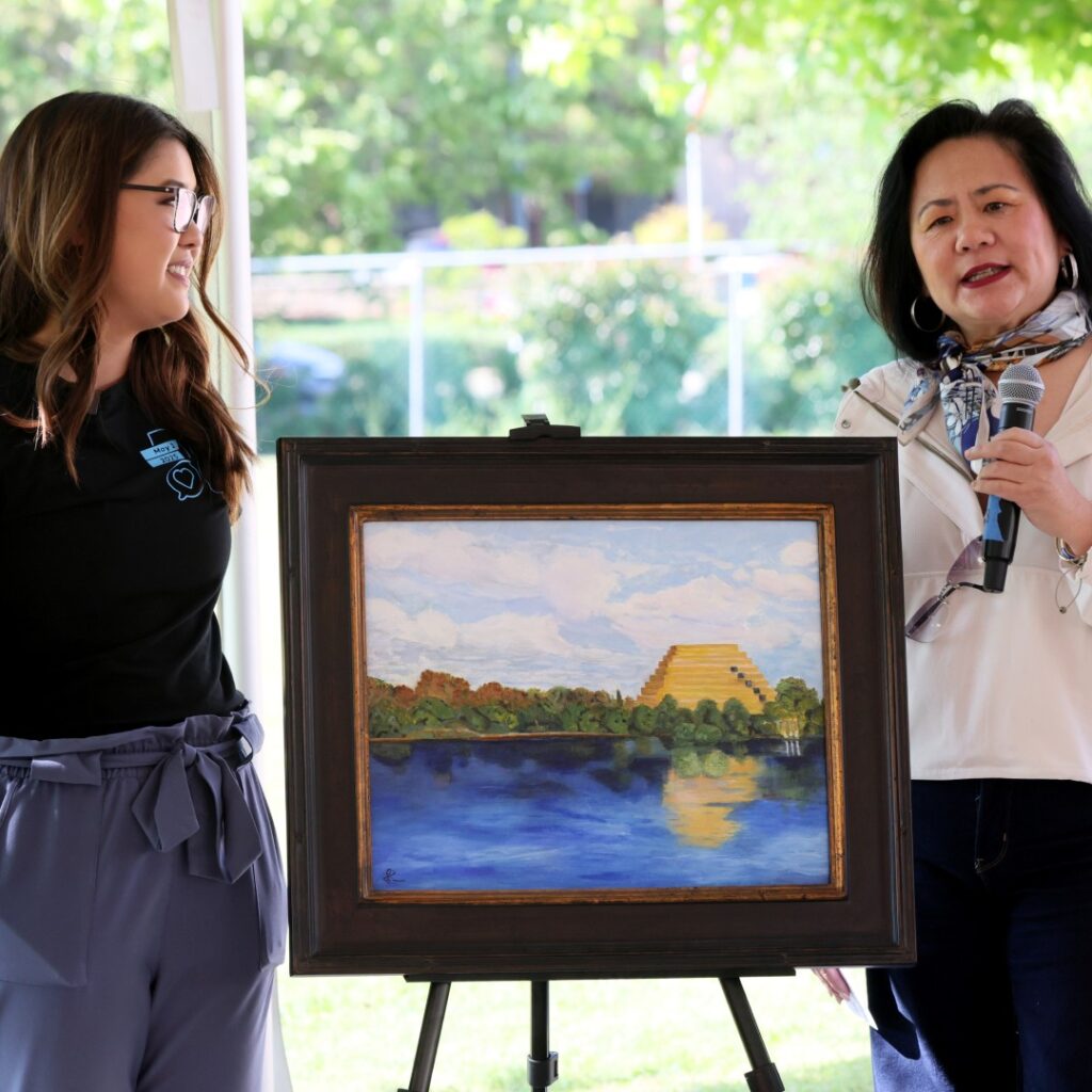 Two women stand beside a painting on an easel outdoors. One woman holds a microphone while speaking; the other smiles at her. The painting depicts a yellow pyramid by a lake, with trees and a blue sky reflected in the water.
