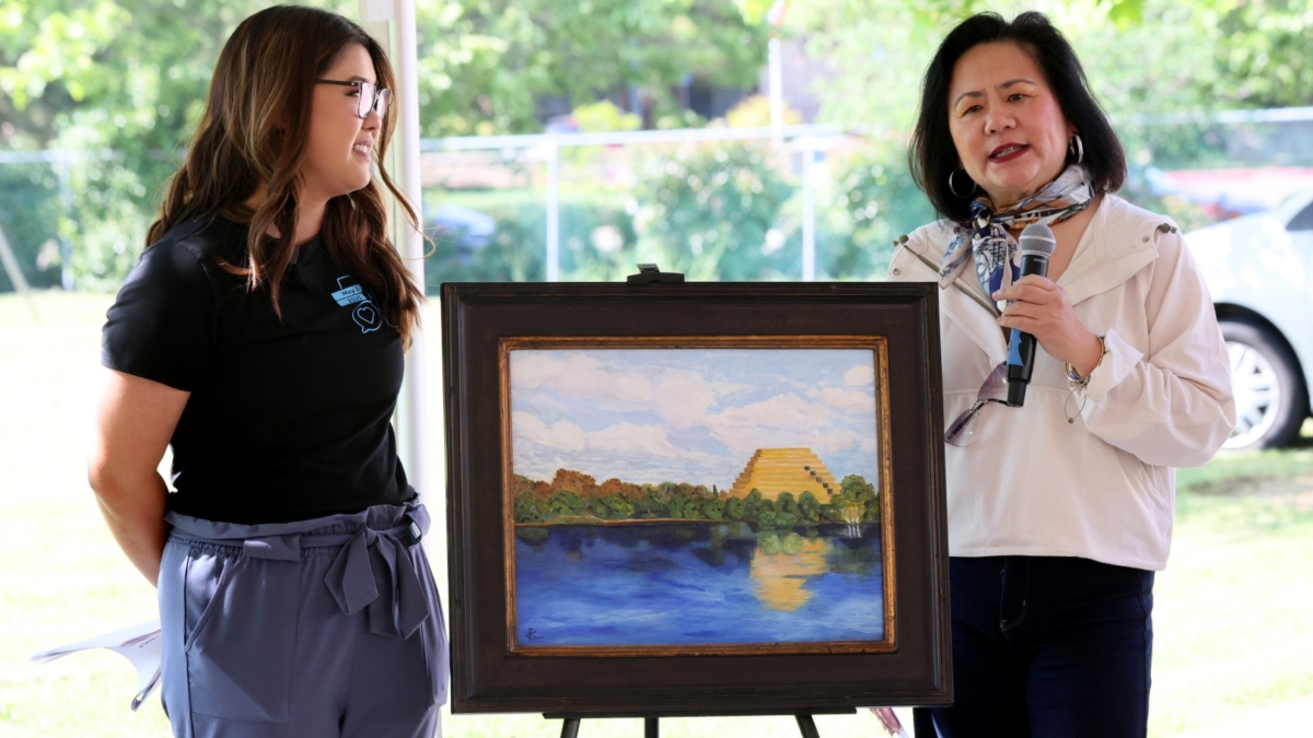 Two women stand beside a painting on an easel outdoors. One woman holds a microphone while speaking; the other smiles at her. The painting depicts a yellow pyramid by a lake, with trees and a blue sky reflected in the water.
