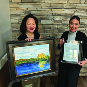 Two women stand indoors in front of a stone wall. One woman holds a framed painting of a riverside landscape; the other holds a clear plaque with text and a small image. Both women are smiling.