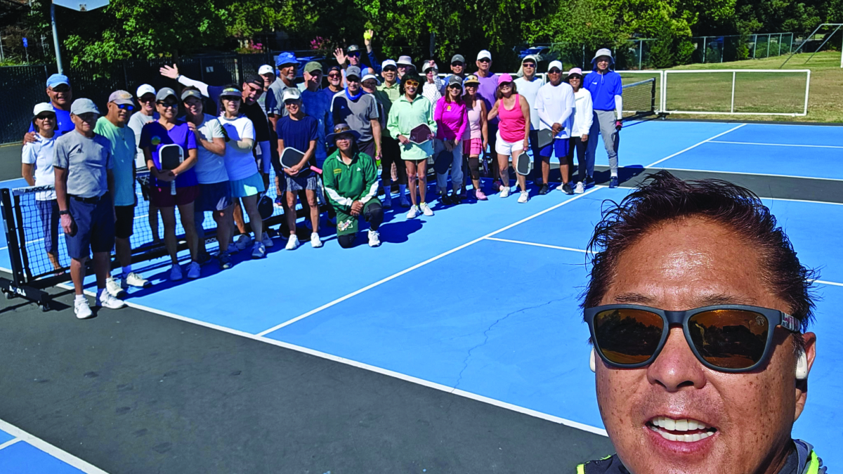 A man in sunglasses takes a selfie in front of a large group of people posing on outdoor tennis courts. Most are holding rackets and smiling under bright sunlight with trees in the background.