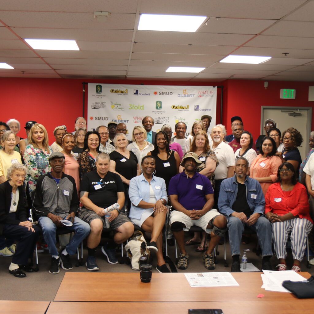 A large group of adults pose together in a conference room. Most are standing, while some sit in front. A banner with logos is on the wall behind them, and tables with papers and a camera are in the foreground.