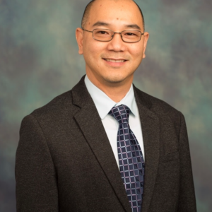A man wearing glasses, a dark suit, a light blue shirt, and a patterned tie is smiling and standing in front of a mottled blue and gray background.