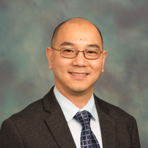 A man wearing glasses, a dark suit, a light blue shirt, and a patterned tie is smiling and standing in front of a mottled blue and gray background.