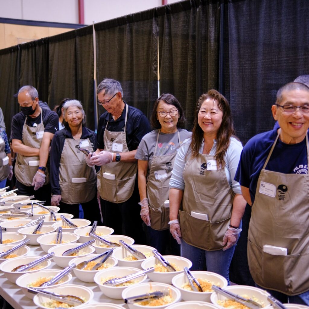 A group of smiling volunteers wearing aprons and gloves stand behind a long table filled with bowls of food, ready to serve at a community event. Black curtains hang behind them.