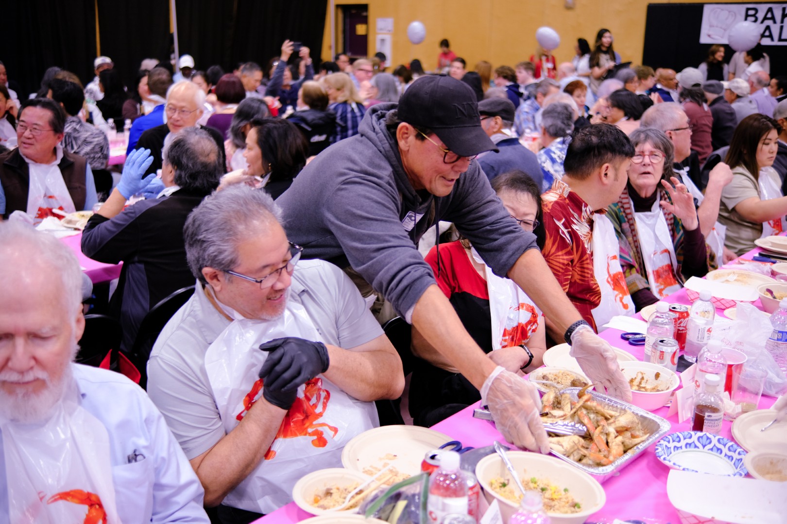 A man wearing a cap serves seafood to smiling people seated at crowded tables covered with food and drinks at a lively indoor community gathering.