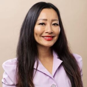 A woman with long dark hair wearing a light purple buttoned blouse is smiling in front of a plain beige background.