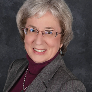 A smiling older woman with short gray hair and glasses, wearing a gray blazer over a maroon turtleneck and a beaded necklace, poses in front of a dark textured background.