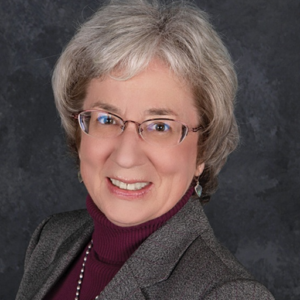A smiling older woman with short gray hair and glasses, wearing a gray blazer over a maroon turtleneck and a beaded necklace, poses in front of a dark textured background.