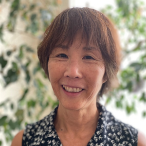 A woman with short brown hair smiles at the camera. She is wearing a dark sleeveless top with a white leaf pattern. The background is softly blurred with green foliage.
