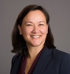 A woman with straight dark hair, wearing a dark blazer and burgundy blouse, smiles at the camera against a plain gray background.