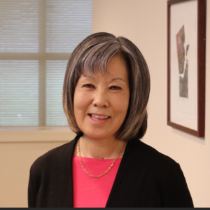 A woman with short gray hair and bangs, wearing a pink top, black cardigan, and gold necklace, smiles while standing indoors near framed art and a window with blinds.