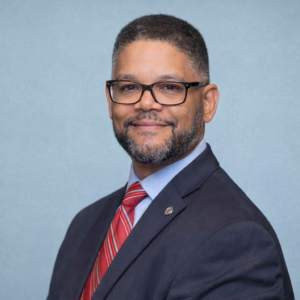 A man with short hair, glasses, and a beard is wearing a dark suit, blue shirt, and red striped tie, smiling at the camera against a plain light blue background.