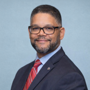 A man with short hair, glasses, and a beard is wearing a dark suit, blue shirt, and red striped tie, smiling at the camera against a plain light blue background.