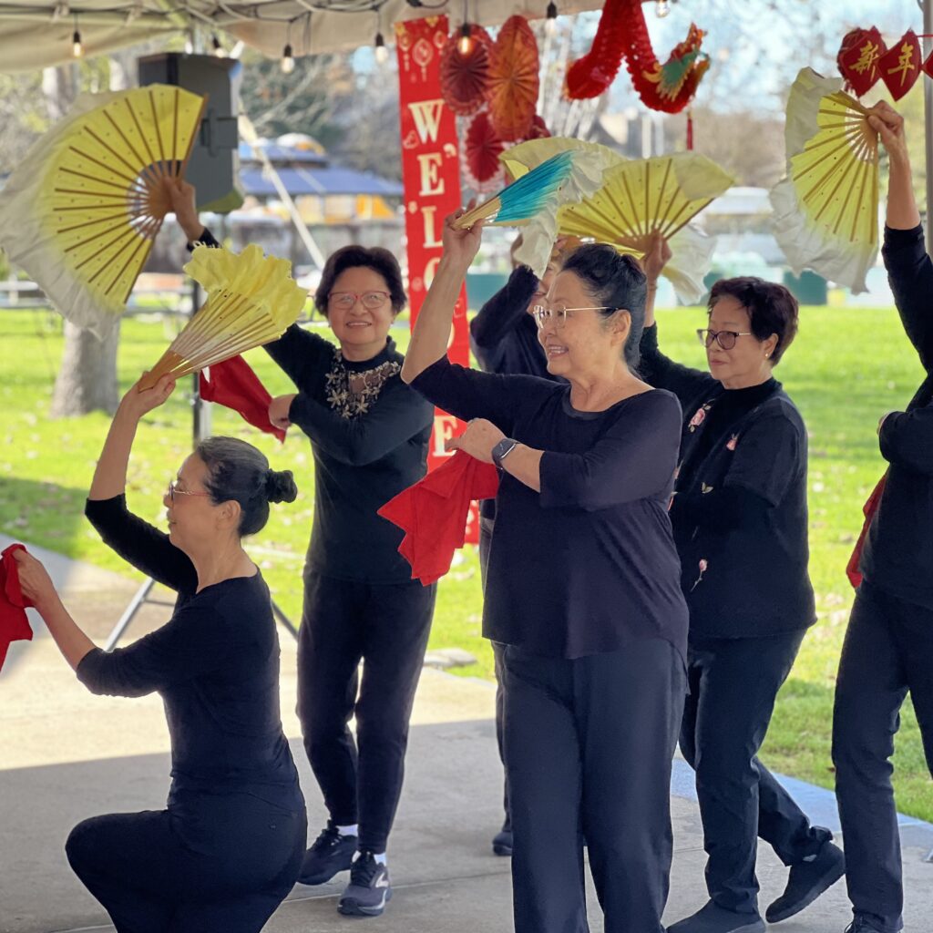 Five women in black outfits perform a fan dance outdoors, holding colorful fans and red cloths. They smile and pose gracefully under festive decorations with trees and grass visible in the background.