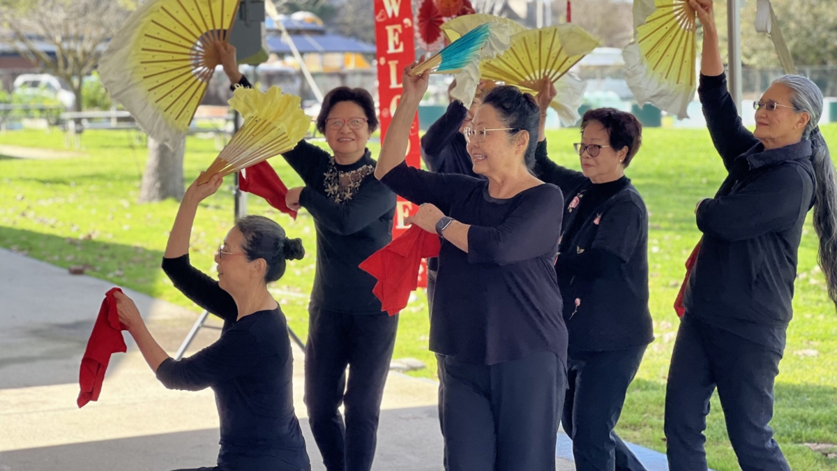 Five women in black outfits perform a fan dance outdoors, holding colorful fans and red cloths. They smile and pose gracefully under festive decorations with trees and grass visible in the background.