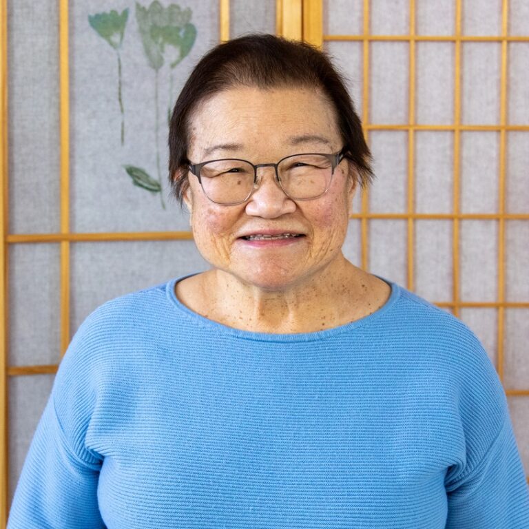An older woman wearing glasses and a light blue sweater smiles while standing in front of a decorative wooden room divider with floral designs.