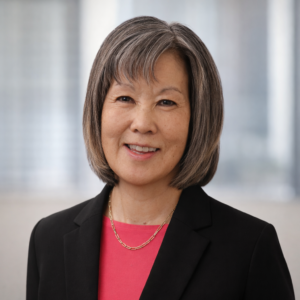 A woman with short gray hair, wearing a black blazer over a pink top and a gold necklace, smiles at the camera. The background is softly blurred with large windows.