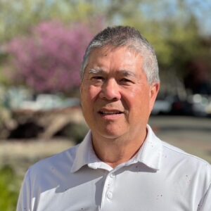 An older man with short gray hair smiles outdoors, wearing a white polo shirt. The background features blurred trees with green and purple foliage on a sunny day.