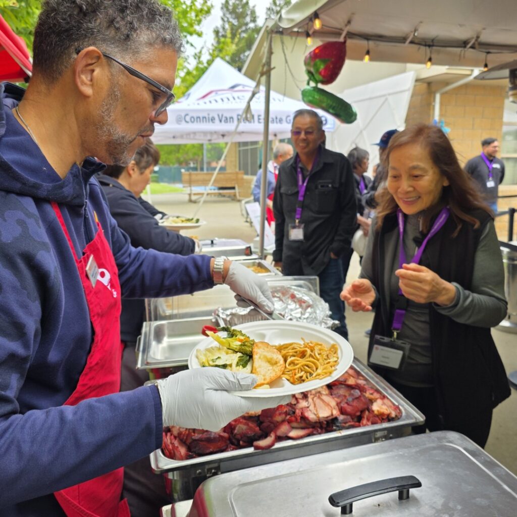 A man wearing an apron and gloves serves food, including noodles and meat, to a smiling woman holding a plate at an outdoor event with people gathered under tents in the background.