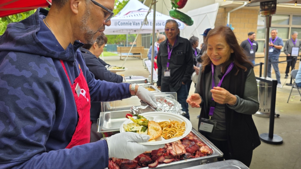 A man wearing an apron and gloves serves food, including noodles and meat, to a smiling woman holding a plate at an outdoor event with people gathered under tents in the background.