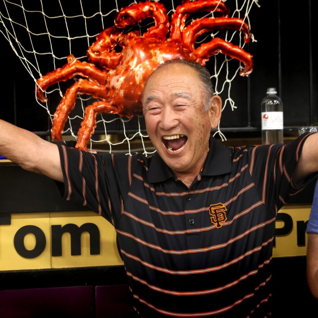 An older man in a black and orange striped shirt smiles joyfully with arms raised; a large decorative crab and fishing net hang behind him, and another person stands partially out of view.