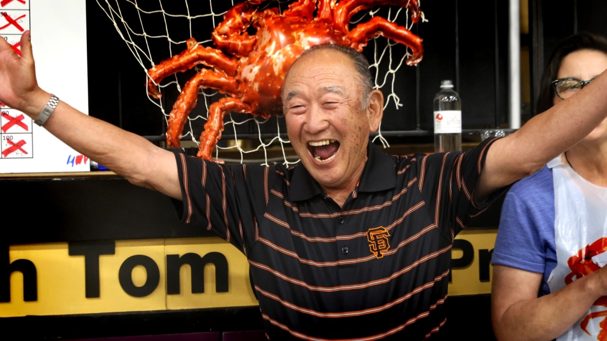 An older man in a black and orange striped shirt smiles joyfully with arms raised; a large decorative crab and fishing net hang behind him, and another person stands partially out of view.