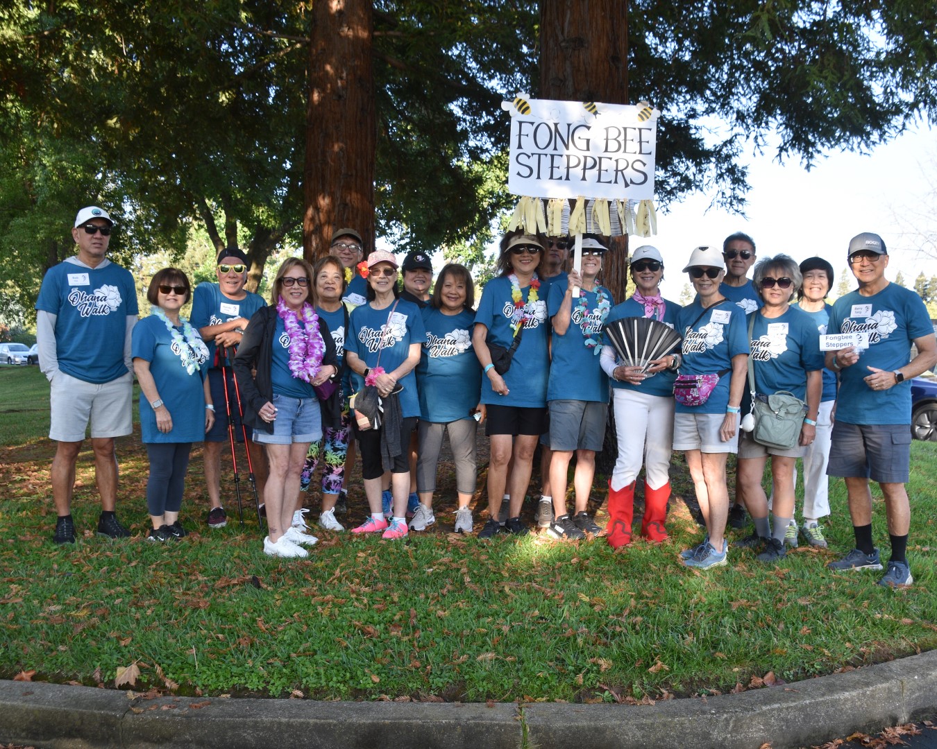 A group of people in blue “Fong Bee Steppers” shirts stand together outdoors on grass, smiling for the camera at the Ohana Walk fundraiser for ACC Senior Services. Two hold a “Fong Bee Steppers” sign; some wear leis or hold fans, with trees in the background.