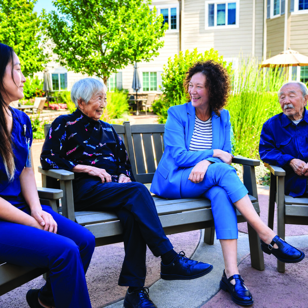 Four people, including two elderly individuals, sit and chat on benches outdoors in a garden, enjoying a sunny day. A woman in blue scrubs and another in a blue suit smile and engage in conversation.