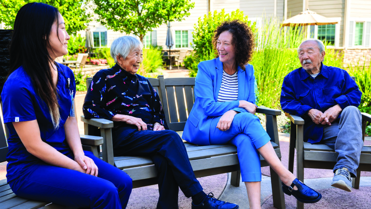 Four people, including two elderly individuals, sit and chat on benches outdoors in a garden, enjoying a sunny day. A woman in blue scrubs and another in a blue suit smile and engage in conversation.