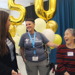 Three women smile and talk at a celebration with yellow balloons and a 50 balloon in the background. One woman wears a name tag reading Julie, and another woman in a striped shirt wears a name tag reading Marcy.