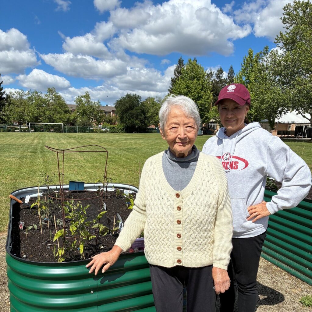 Two women stand by a raised garden bed in a park on a sunny day. The older woman is in front, wearing a cream sweater, and the younger woman behind wears a maroon cap and sweatshirt. Gardening gloves are on the ground.