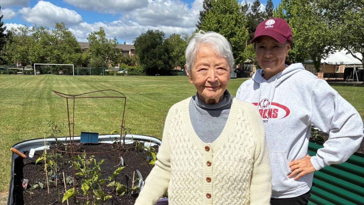 Two women stand by a raised garden bed in a park on a sunny day. The older woman is in front, wearing a cream sweater, and the younger woman behind wears a maroon cap and sweatshirt. Gardening gloves are on the ground.