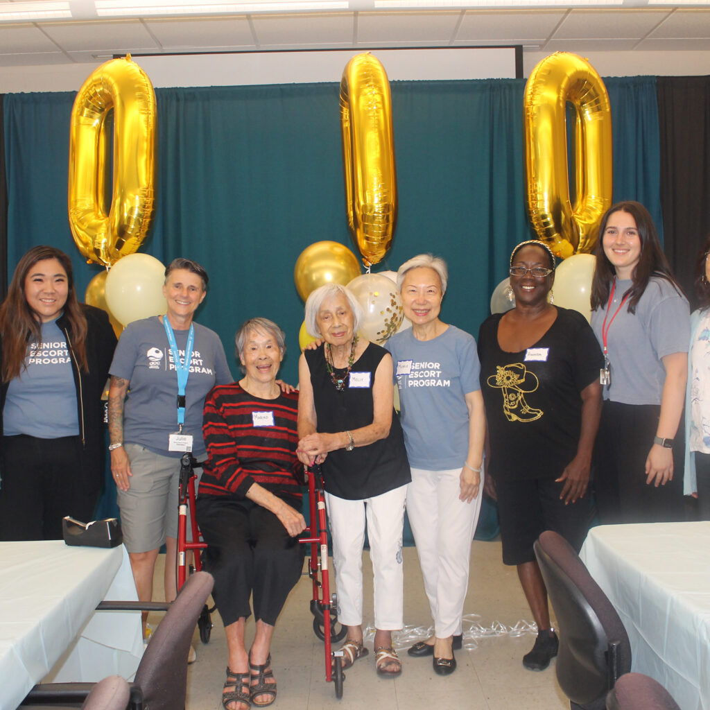 A group of nine people stand smiling in front of large gold 30000 balloons, celebrating at an indoor event with decorated tables and balloons.
